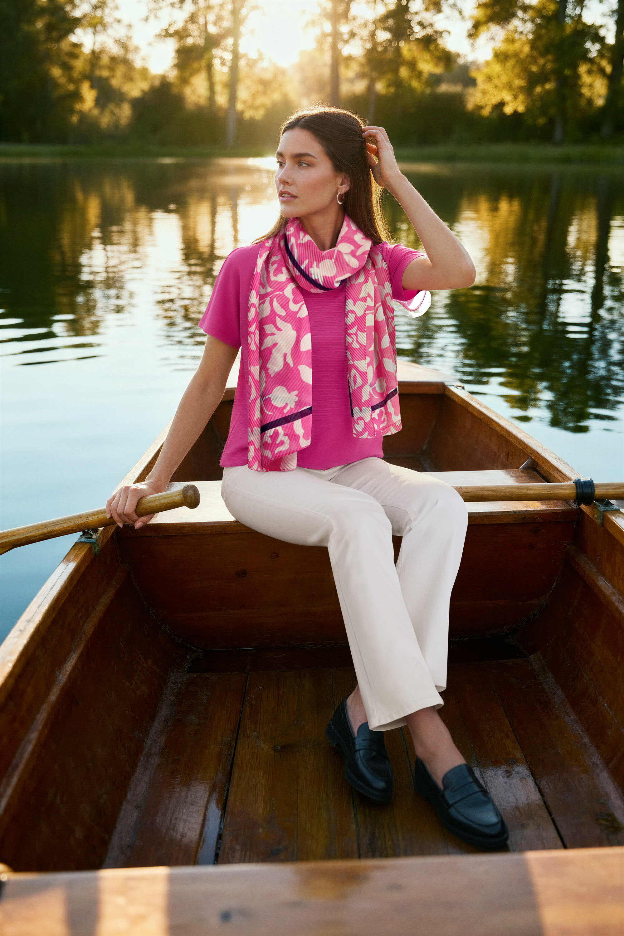 Nő ül egy csónakban, rózsaszín blúzt, krémszínű Bootcut Jeans ‒ Fit »Lou« farmert, bőr mokaszint és Női fülbevaló, Wave fülbevalót visel.