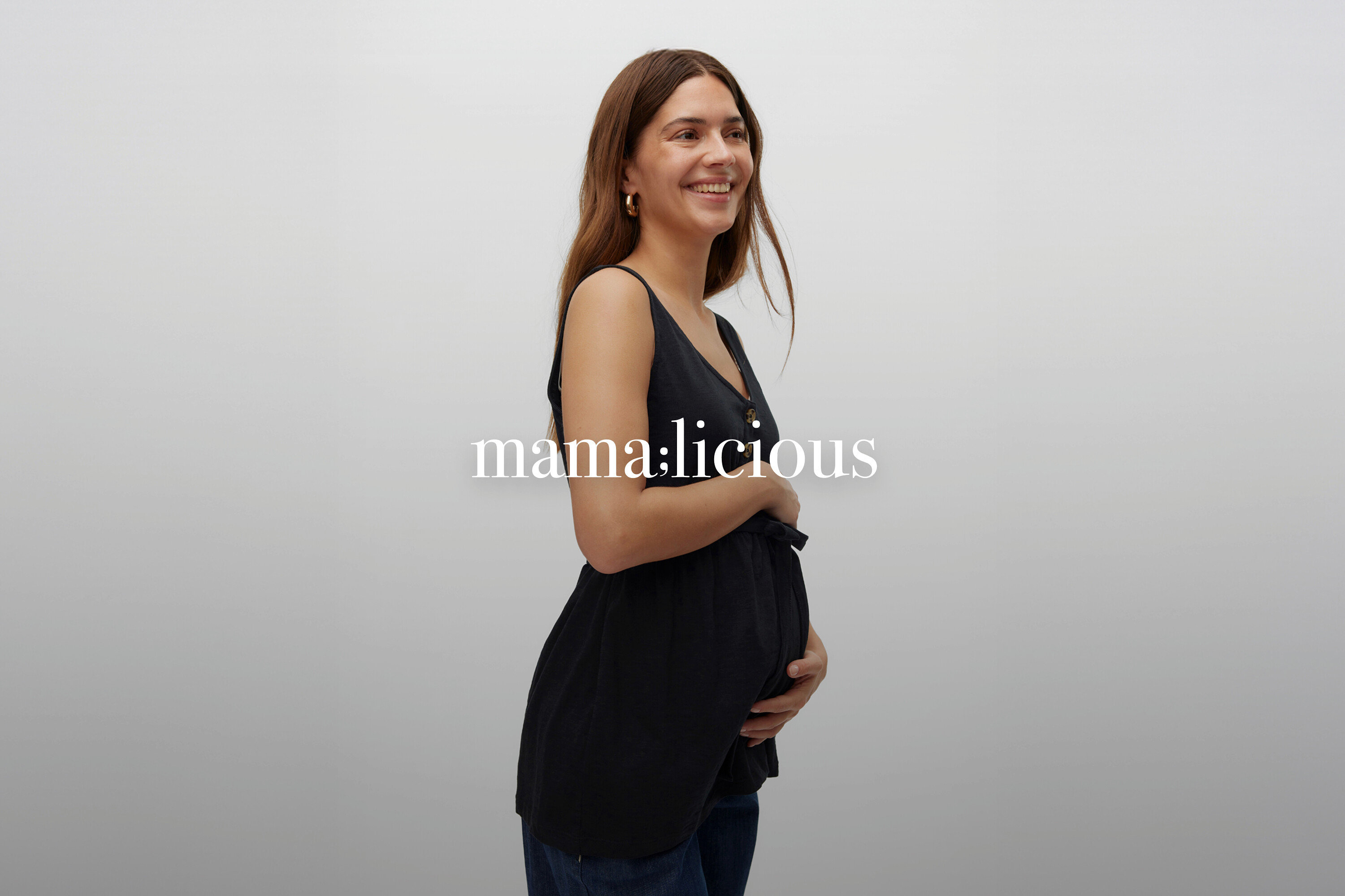 Mosolygó, hosszú barna hajú terhes nő fekete, ujjatlan, gombos kismama felsőben és farmerben.