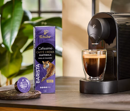 A kávéfőző Cafissimo BARISTA Caffè Crema Guatemala kávét készít egy pohárba. Mellette egy 10 db-os kapszula csomag és egy kapszula látható.