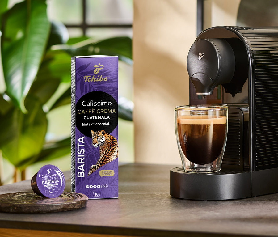 A kávéfőző Cafissimo BARISTA Caffè Crema Guatemala kávét készít egy pohárba. Mellette egy 10 db-os kapszula csomag és egy kapszula látható.
