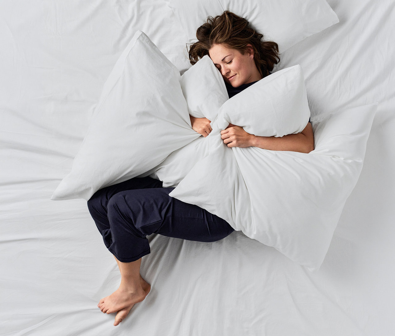 Fehér ágyon alvó nő, nagy fehér párnát ölel. Sötétkék CALIDA »Deepsleepwear Balancing« nadrágot visel.