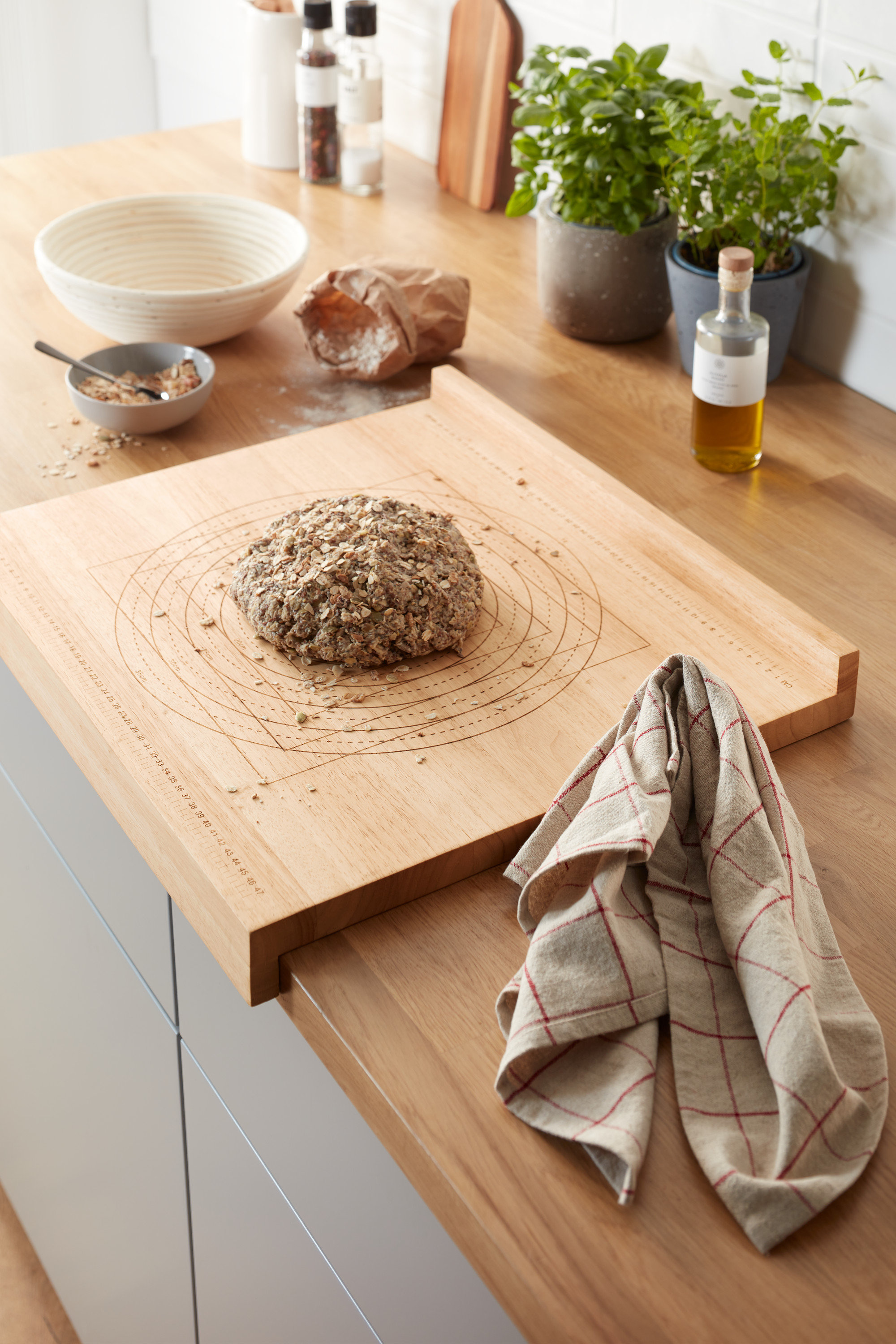 A konyhapulton egy fából készült nyújtódeszka méretskálával, egy kenyérrel. A háttérben cserepes fűszernövények és egyéb konyhai eszközök láthatók.
