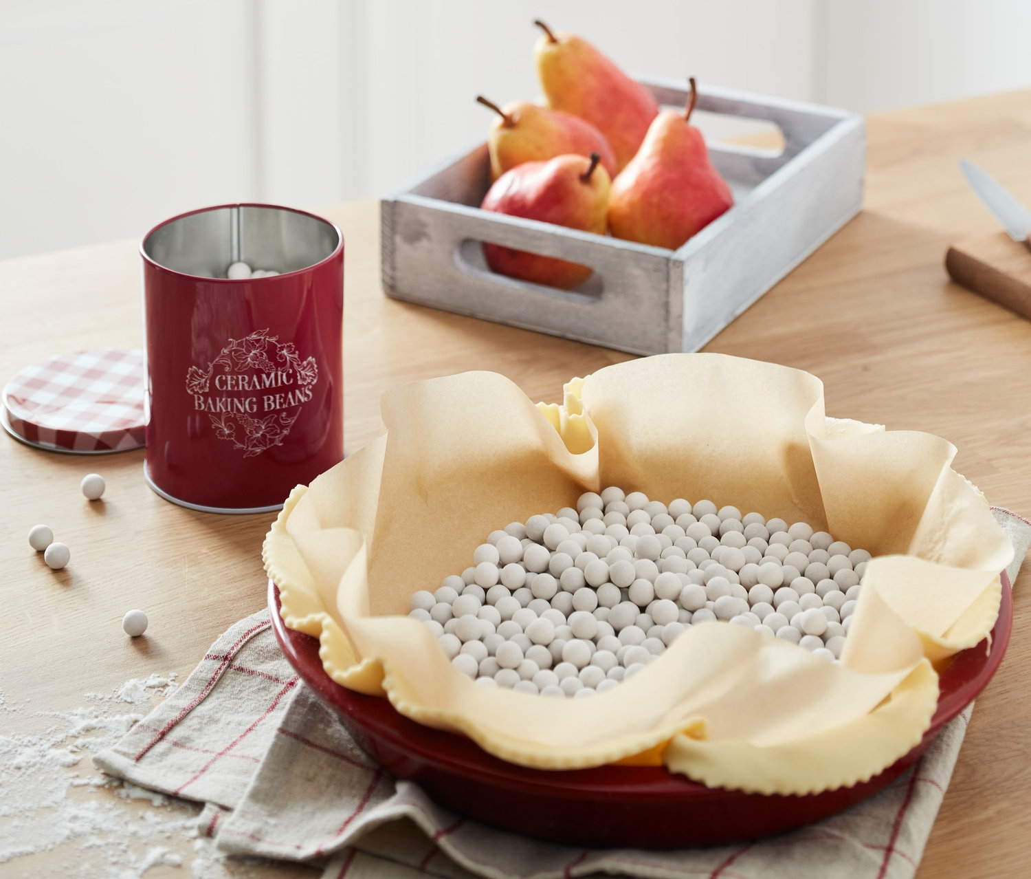 Egy piros piteforma tésztával és kerámia sütőbabokkal. Egy doboz kerámia sütőbab és egy láda körte egy fa asztalon.