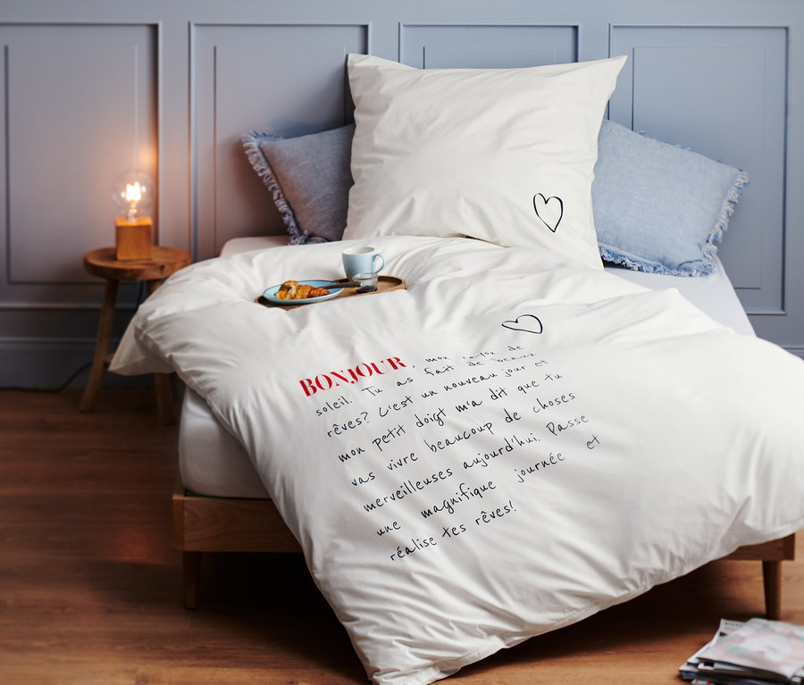 Fehér, normál méretű prémium pamut ágynemű Bonjour felirattal és egy kis szívvel a párnán egy ágyon.