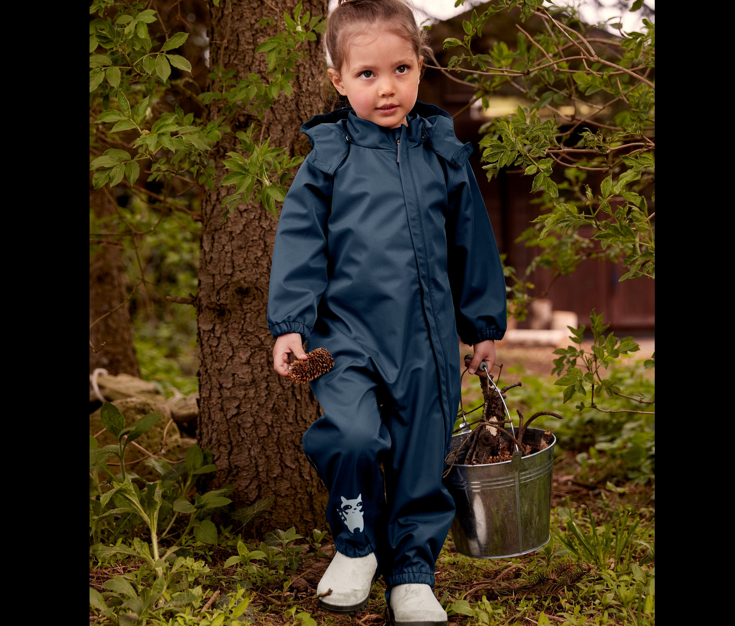 Kislány áll a fa mellett fehér gyerek gumicsizmában és sötétkék kisgyerek esőoverálban, kezében tobozt és vödröt tart ágakkal.