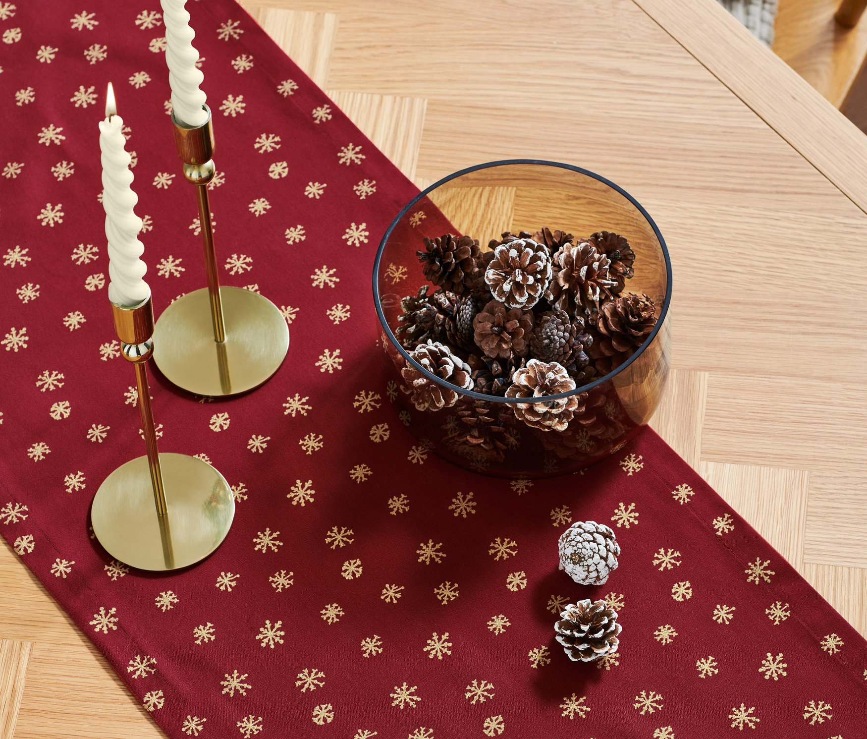 Egy fa asztalon egy piros, arany hópelyhes asztali futó látható, rajta két arany gyertyatartó fehér gyertyákkal és egy üvegtál tele tobozokkal.
