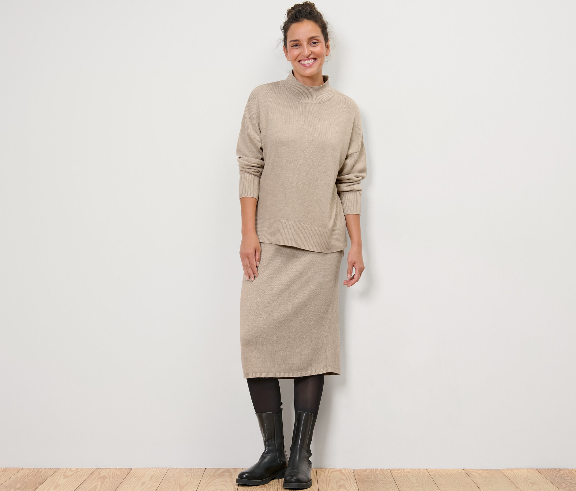 Egy nő áll, finomkötésű pulóvert és kötött szoknyát visel.