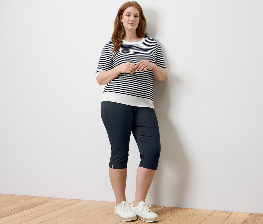 Nő pózol egy finom kötésű csíkos pulóverben és egy sötétkék 3/4-es sztreccsnadrágban.
