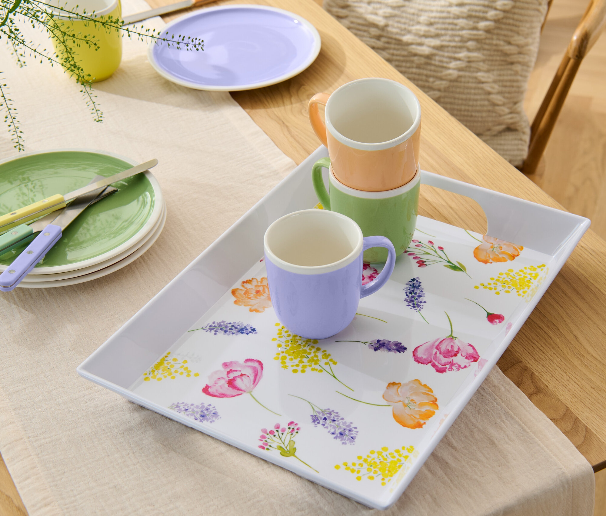 Az asztalon tányérok és kések állnak egy virágos tálca mellett, amelyen három színes bögre áll.