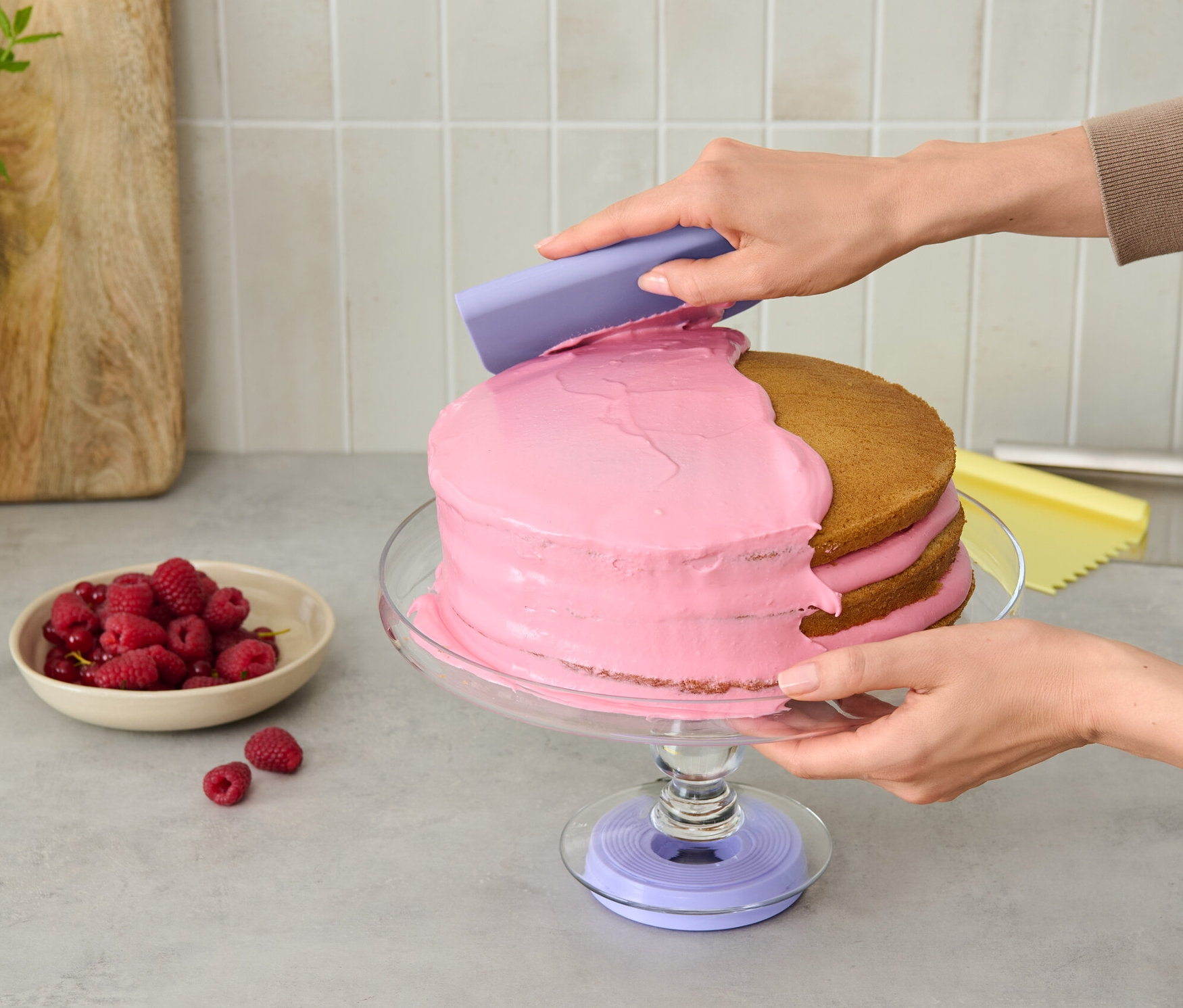 Személy rózsaszín krémet visz fel egy tortára egy lila eszközzel, amely egy forgatható tortatartó talpon áll.