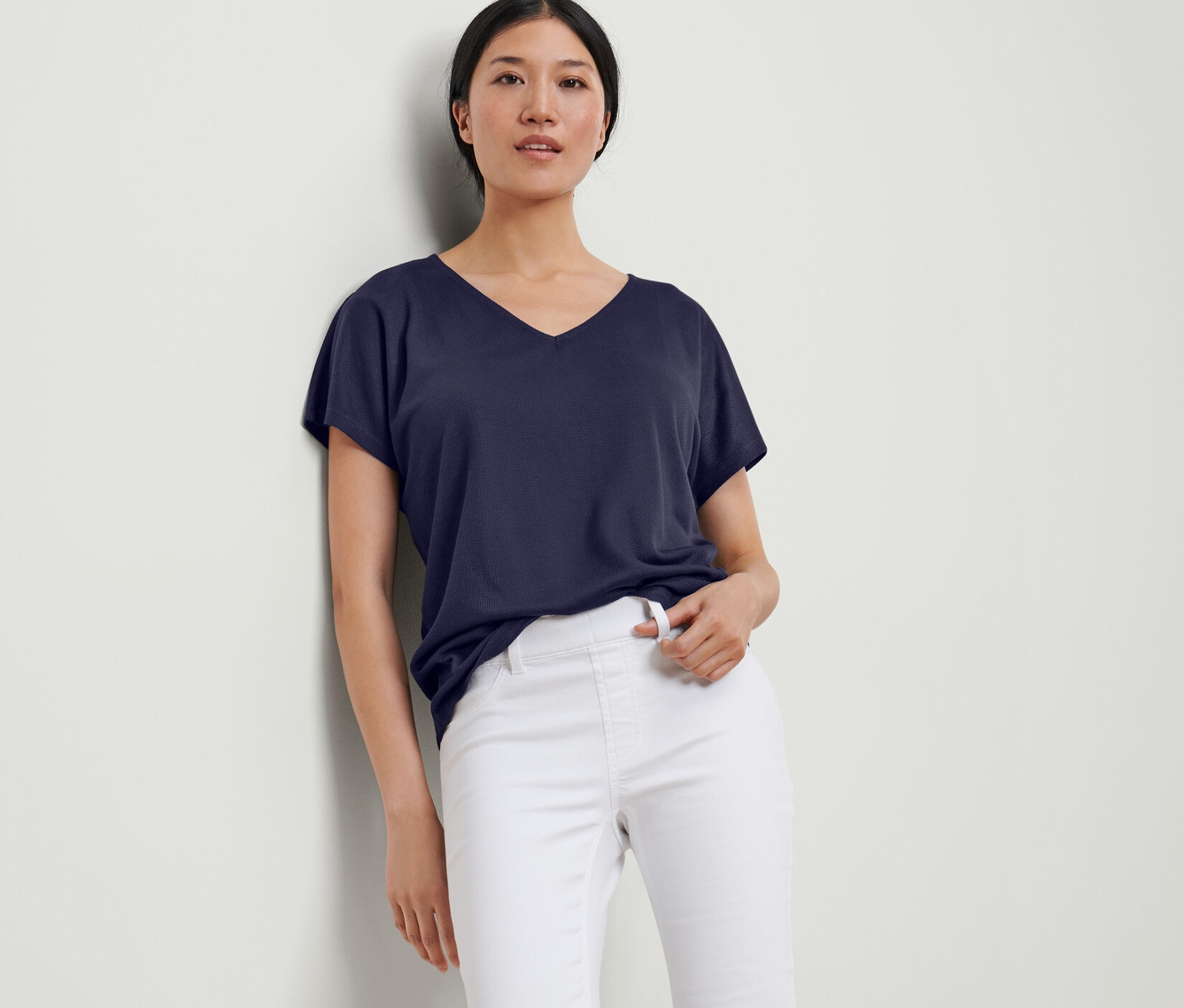 Egy nő a falnak dőlve sötétkék, strukturált felsőt és fehér nadrágot visel.