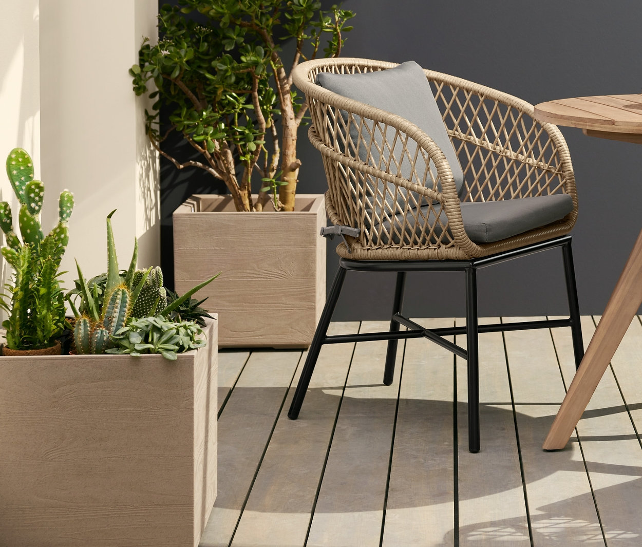 Két polyrattan kerti étkezőszék egy kerek kerti asztalnál áll. Az asztal fölött napelemes függőlámpa lóg. Balra gurulós virágtartó kaspó látható.