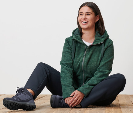 Nő ül a fa padlón, melírozott zöld kapucnis kötött polárfelsőt és termonadrágot visel.
