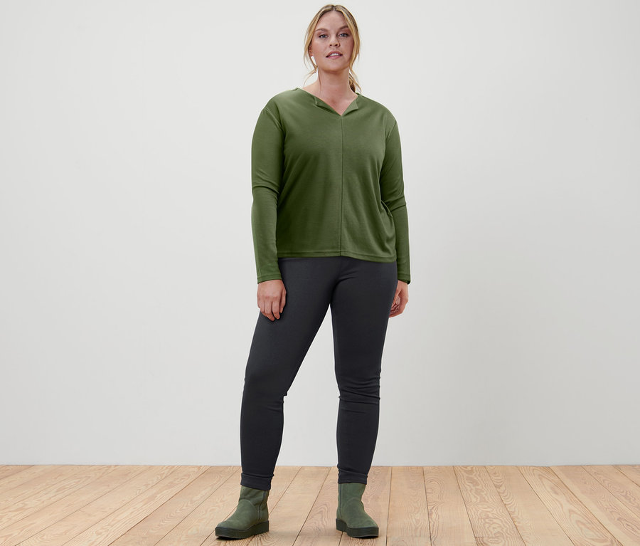Egy nő áll, és női hosszú ujjú pólóblúzt, V-nyakút, olívazöld, női jeggingst, fekete és női velúrbőr csizmát, zöld színt visel.