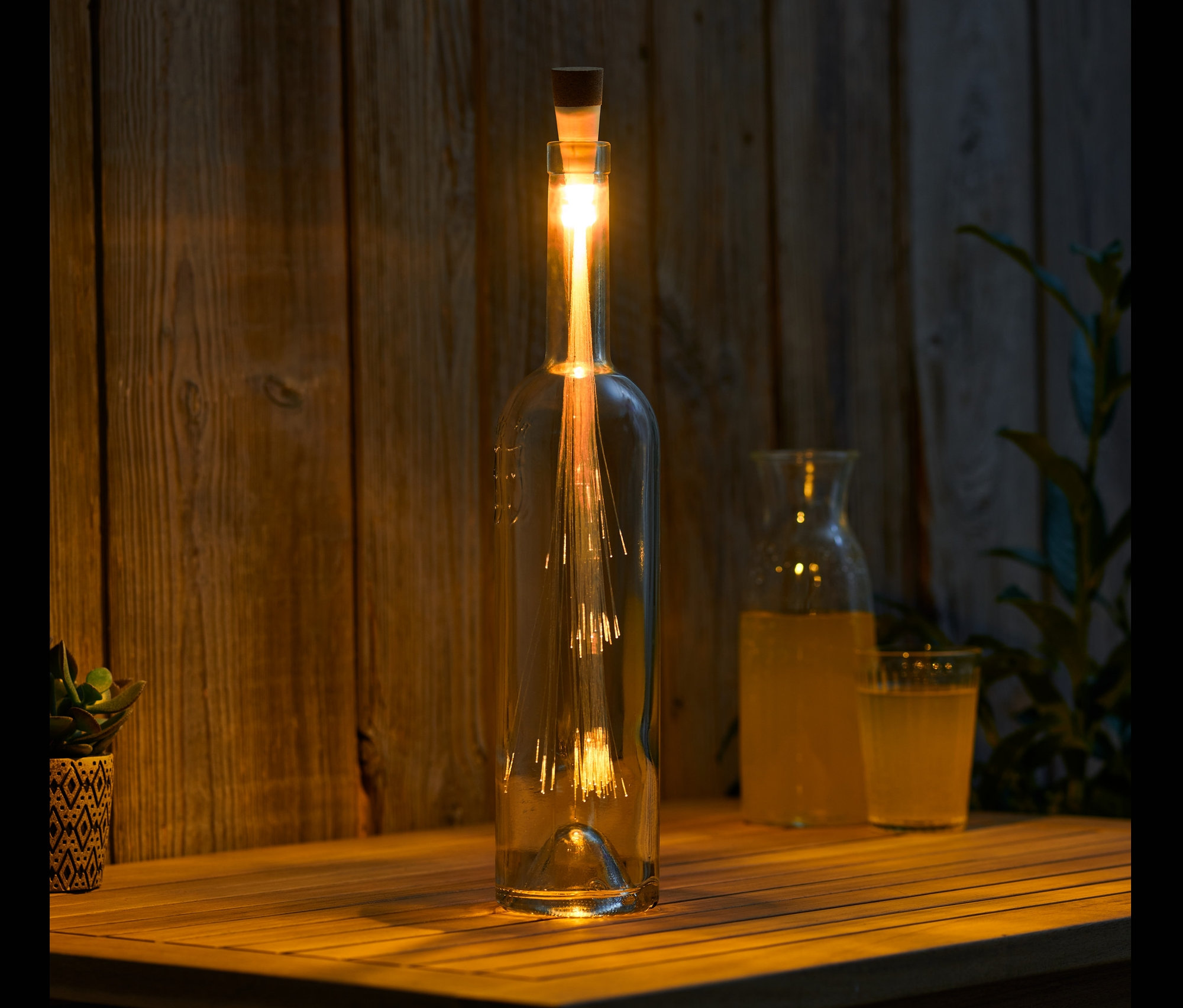 Egy fa asztalon egy üveg áll LED-es dugó akkumulátorral, mellette egy kancsó és egy pohár ital.