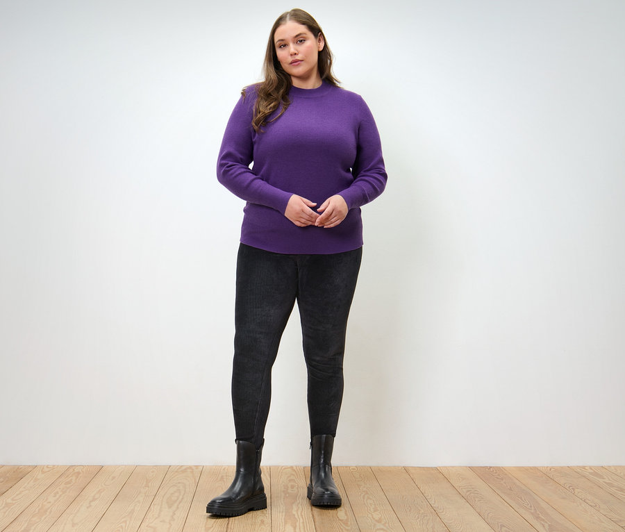 Nő pózol lila finomkötésű pulóverben és fekete kordbársony leggingsben.