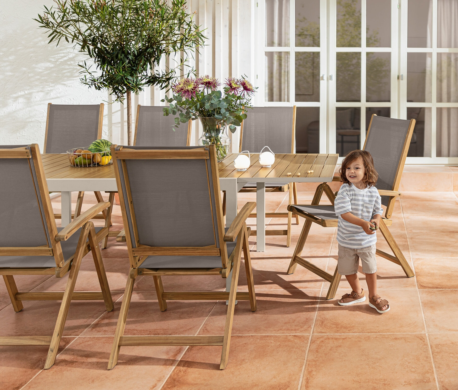 Egy fiú áll egy »Liska« kihúzható alumínium asztal és egy »Lenja« fa magas támlás szék mellett. A háttérben egy zsúrkocsi és egy akkumulátoros asztali lámpa szett látható.