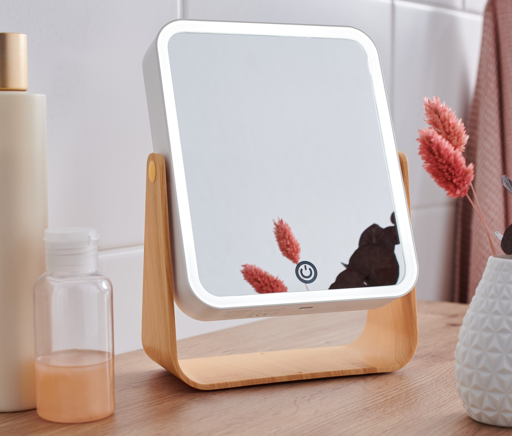 Egy LED-es kozmetikai tükör áll egy faasztalon, üvegek és egy virágokkal teli váza mellett.