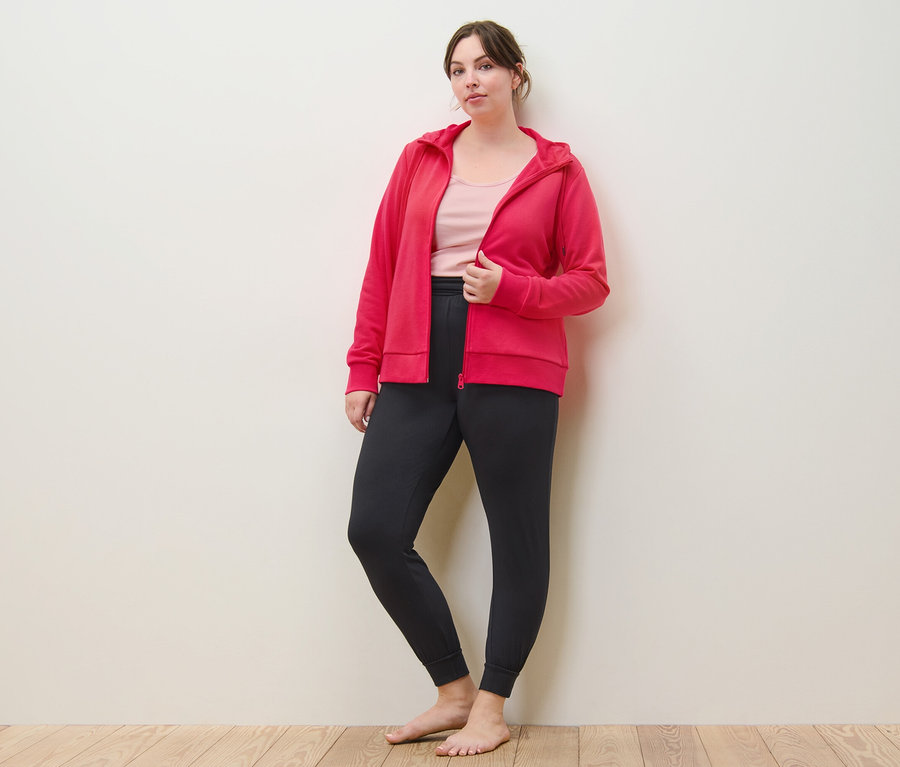 Nő pózol rózsaszín kapucnis szabadidőfelsőben, jóganadrágban és 2 rózsaszín/pink topban.