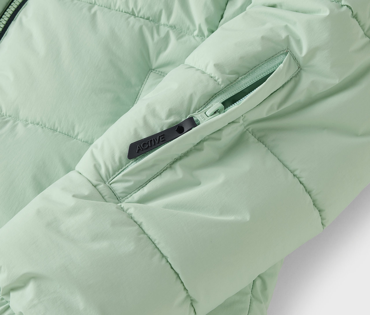 Egy világoszöld, steppelt kabát részlete, cipzáras zsebbel.