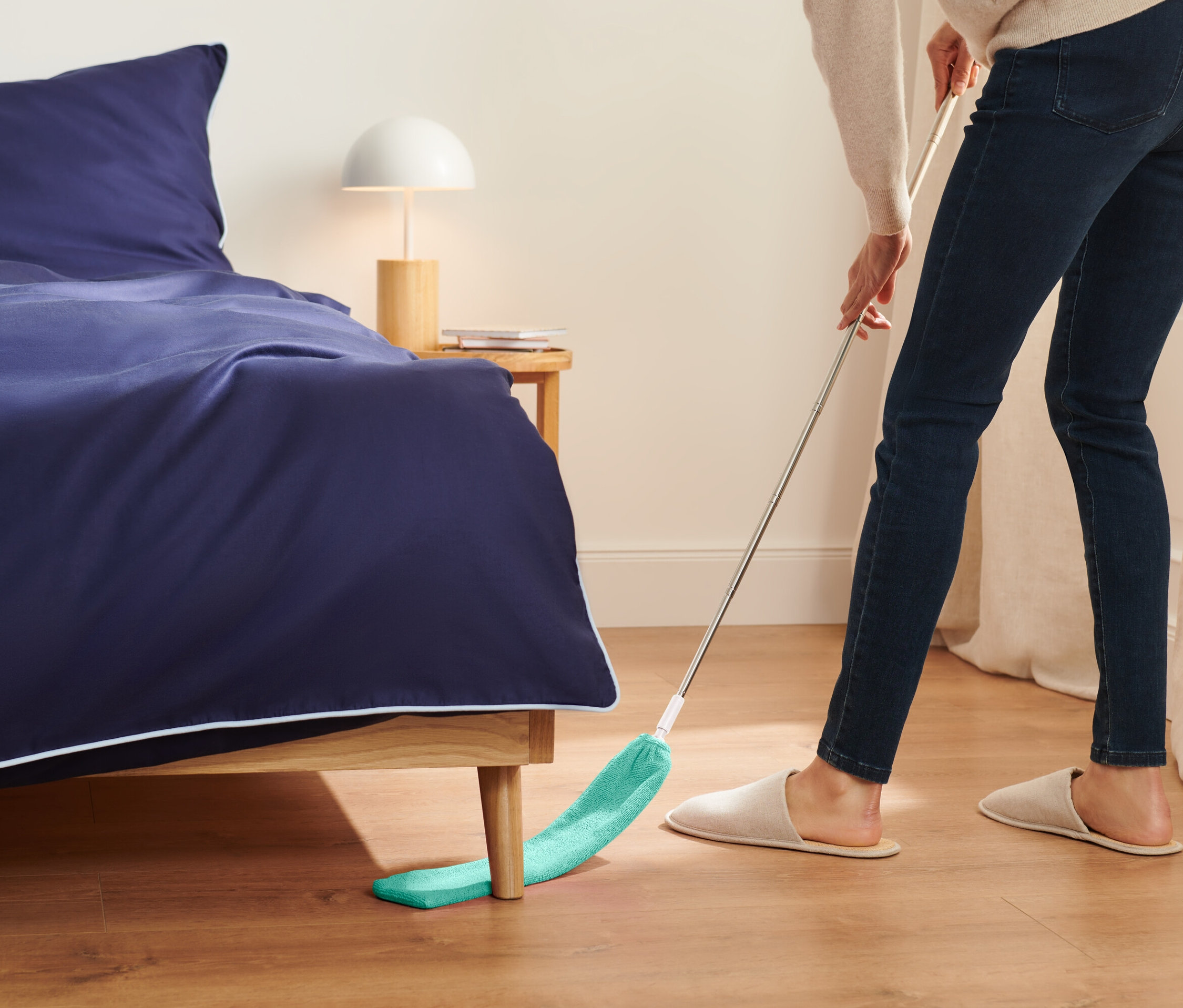 Egy nő zöld felmosóval takarítja a padlót az ágy alatt.