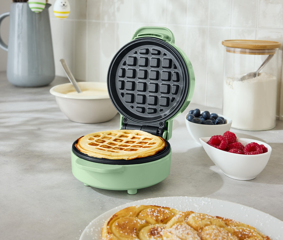 Egy nyitott, kerek, mentazöld gofrisütő, benne egy gofri, egy konyhapulton áll. Mellette egy tányéron két spirális sütemény, egy tál áfonya és egy tál málna található.