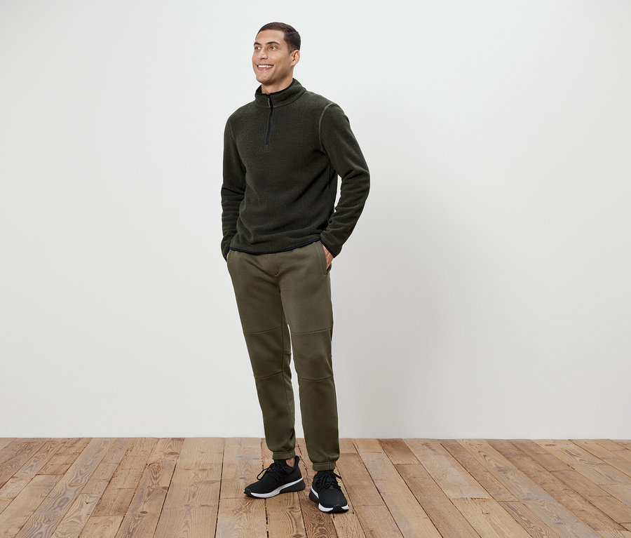 Olívazöld nadrágot és sötétszürke cipzáras gyapjú pulóvert viselő férfi áll a fapadlón.