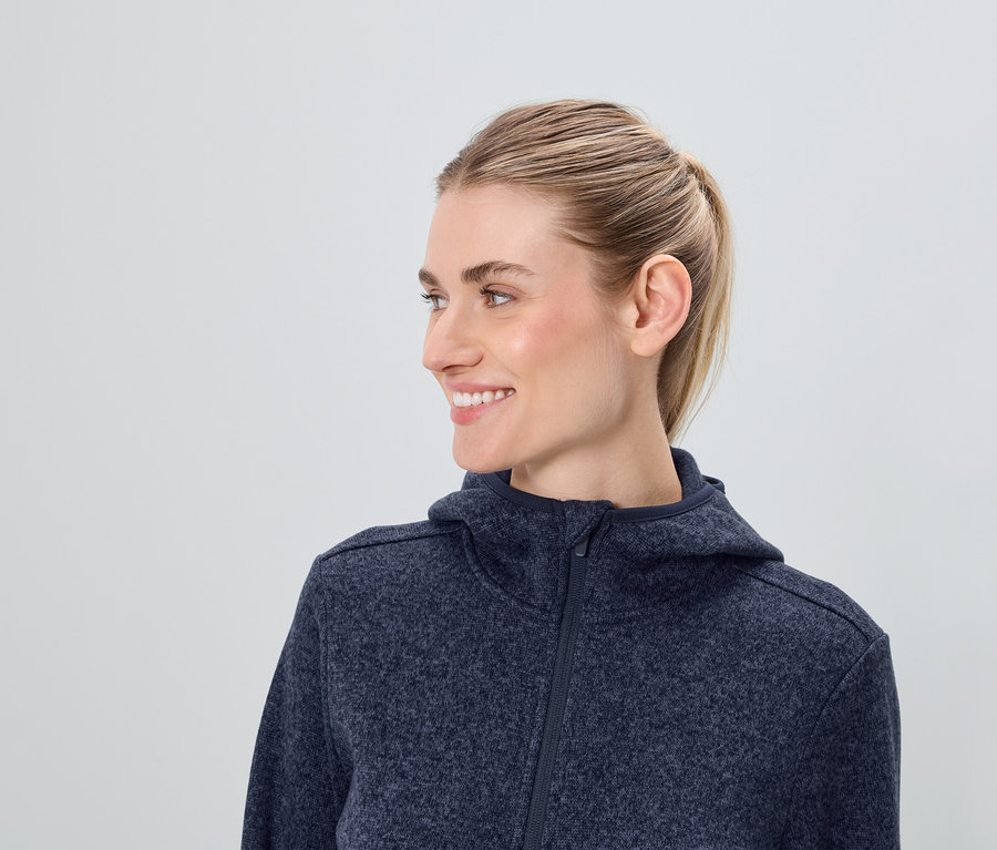 Egy mosolygó, szőke hajú nő profilja kék, cipzáras kapucnis pulóverben.