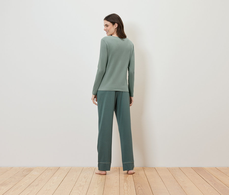 Egy nő áll a kamerának háttal, zöld, hosszú ujjú pizsamában és mintás nadrágban.