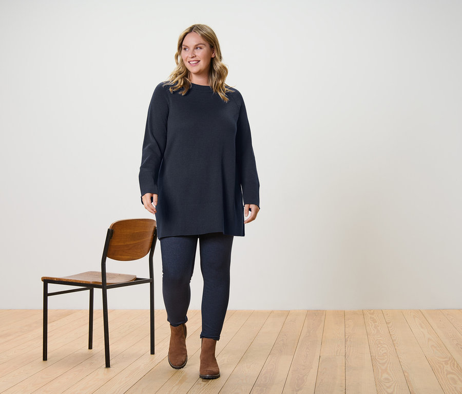 Egy nő sétál, sötétbarna, hosszú kötött pulóvert és farmerhatású, bélelt leggingset visel.
