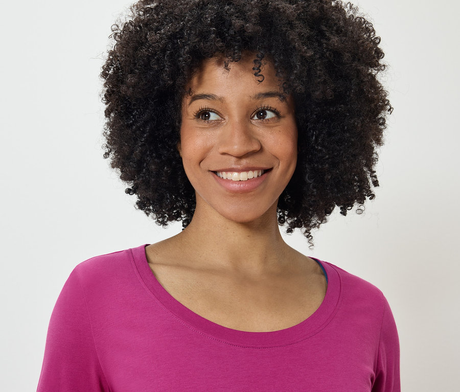 Portré egy mosolygó, afro frizurás nő rózsaszín pólóban, aki balra néz.