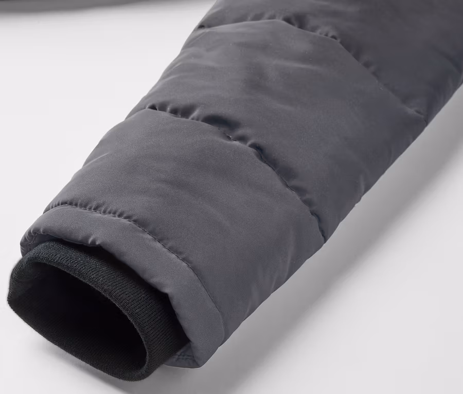 Egy szürke, fényvisszaverő elemekkel ellátott steppelt gyerekkabát részlete, a fekete mandzsettás ujj látható.