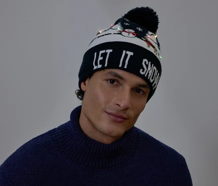 Portré egy férfiról, aki LET IT SNOW feliratú és kis fényekkel díszített karácsonyi sapkát visel.