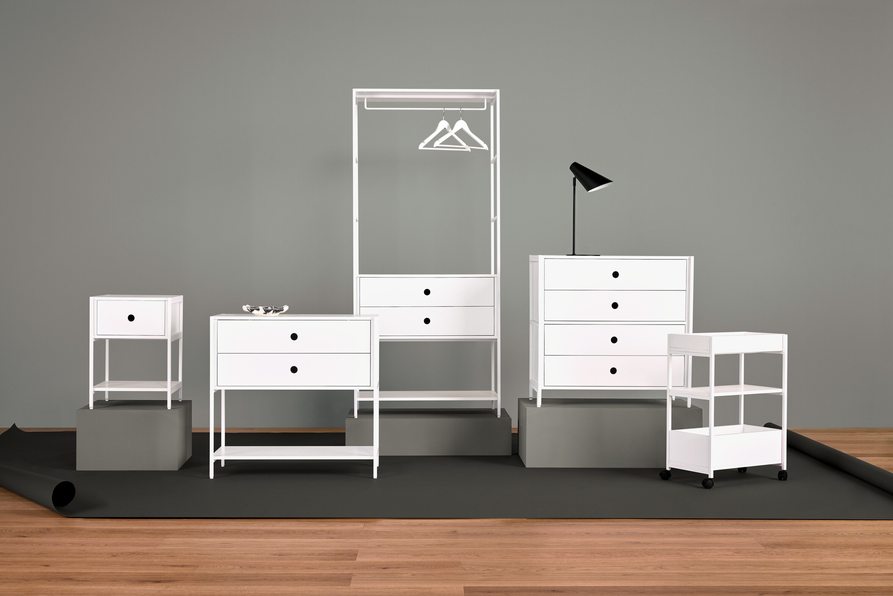 Fehér bútorok elrendezése éjjeliszekrénnyel, fiókos szekrénnyel, ruhaakasztóval, lámpával és polcokkal.