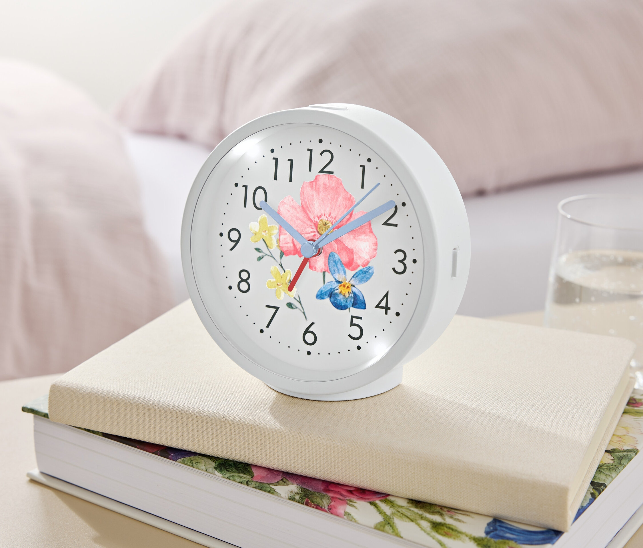 Egy virágmintás fehér ébresztőóra könyveken fekszik egy pohár víz mellett.