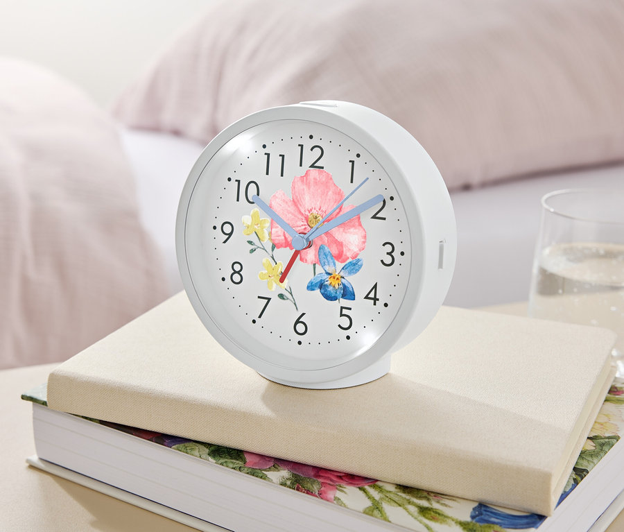 Egy virágmintás fehér ébresztőóra könyveken fekszik egy pohár víz mellett.