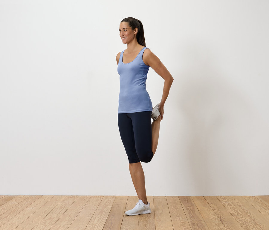Nő kék trikóban és sötét leggingsben nyújtja a combfeszítőjét, miközben a lábát hátul tartja.