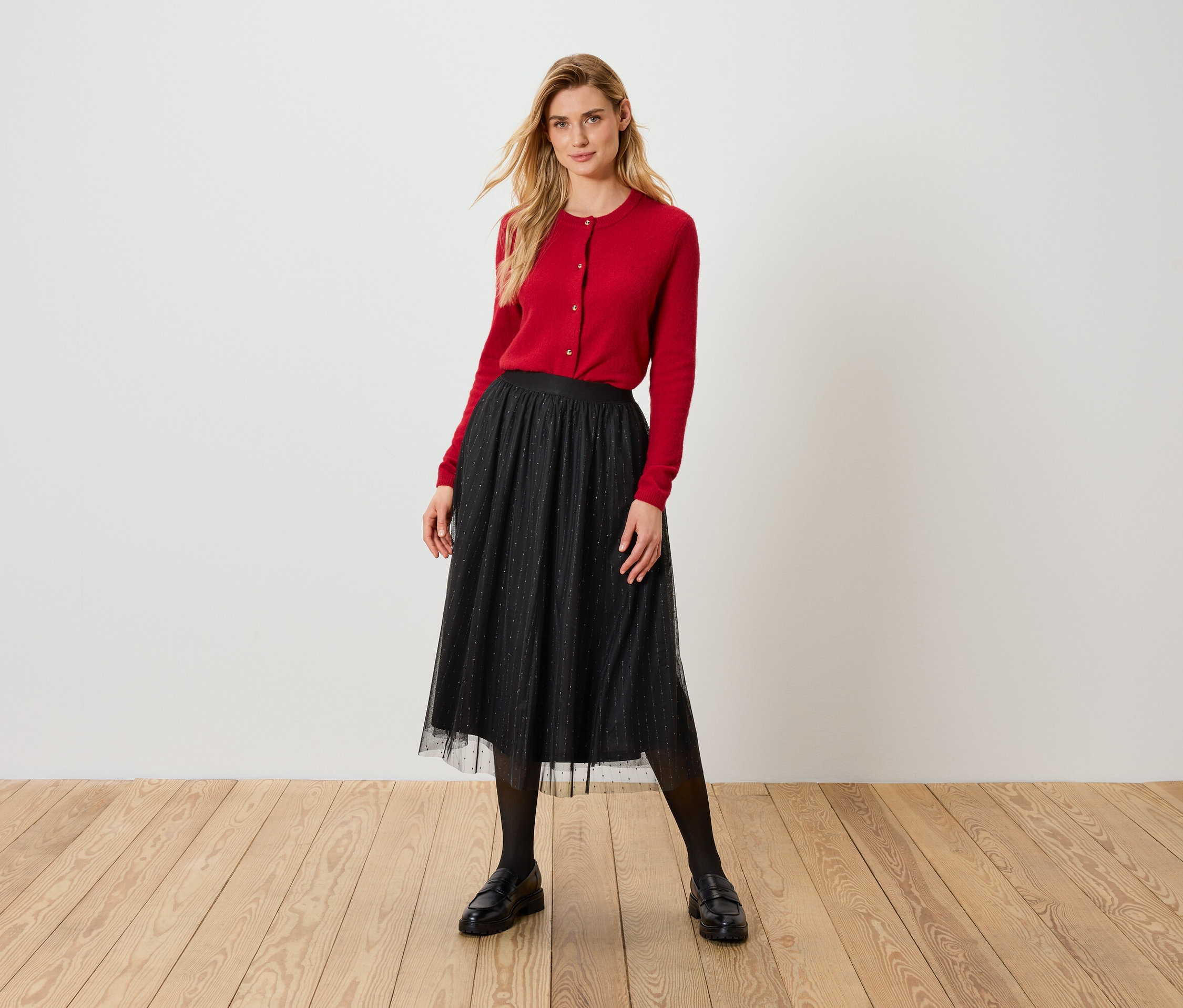 Szőke nő piros kardigánban és fekete tüllszoknyában áll a fapadlón.