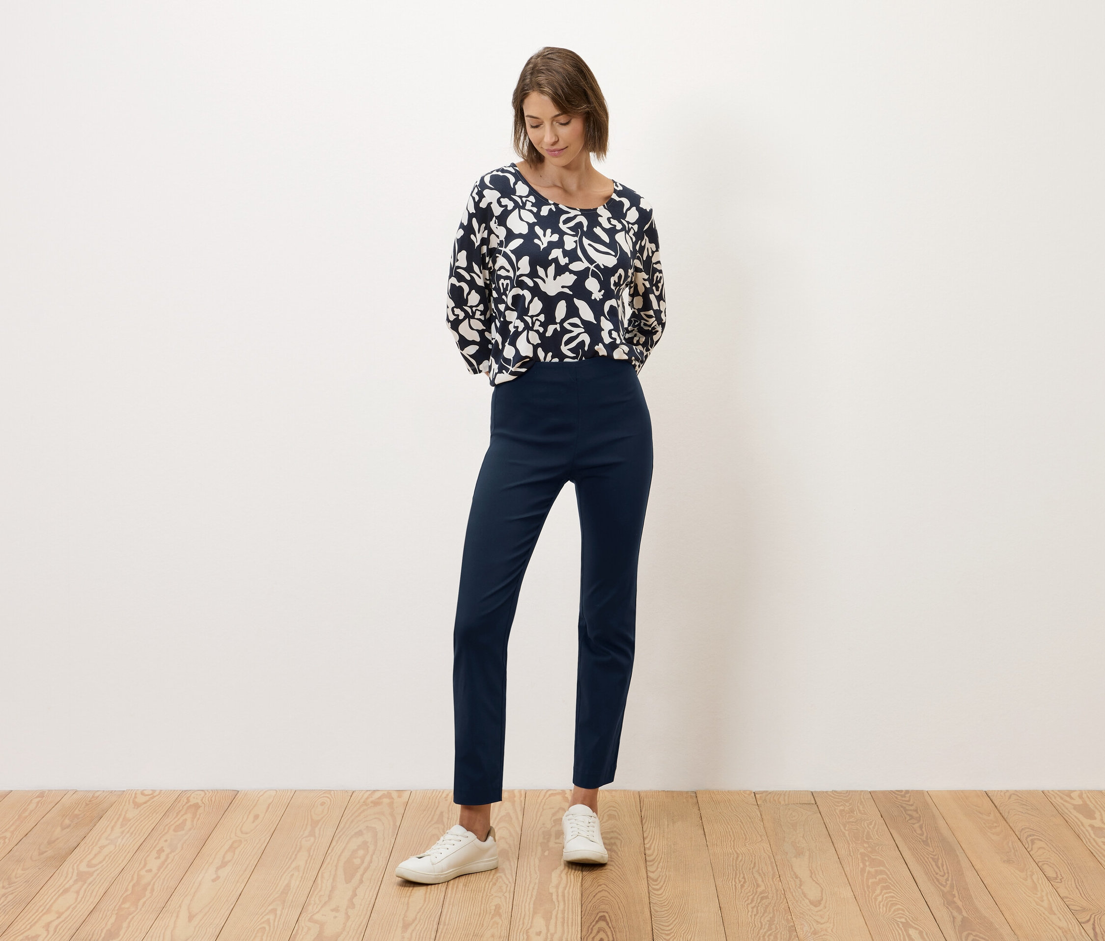 Egy nő áll egy fehér fal előtt, sötétkék nadrágot és fekete-fehér mintás blúzt visel.