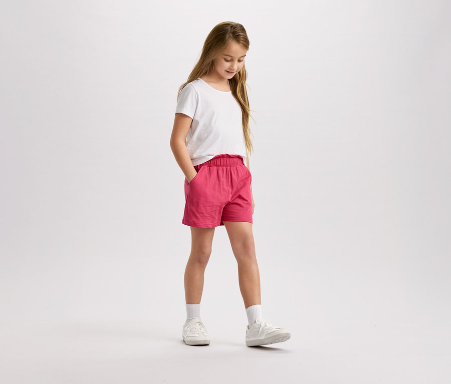 Egy szőke hajú lány fehér pólóban és rózsaszín rövidnadrágban áll és lefelé néz.