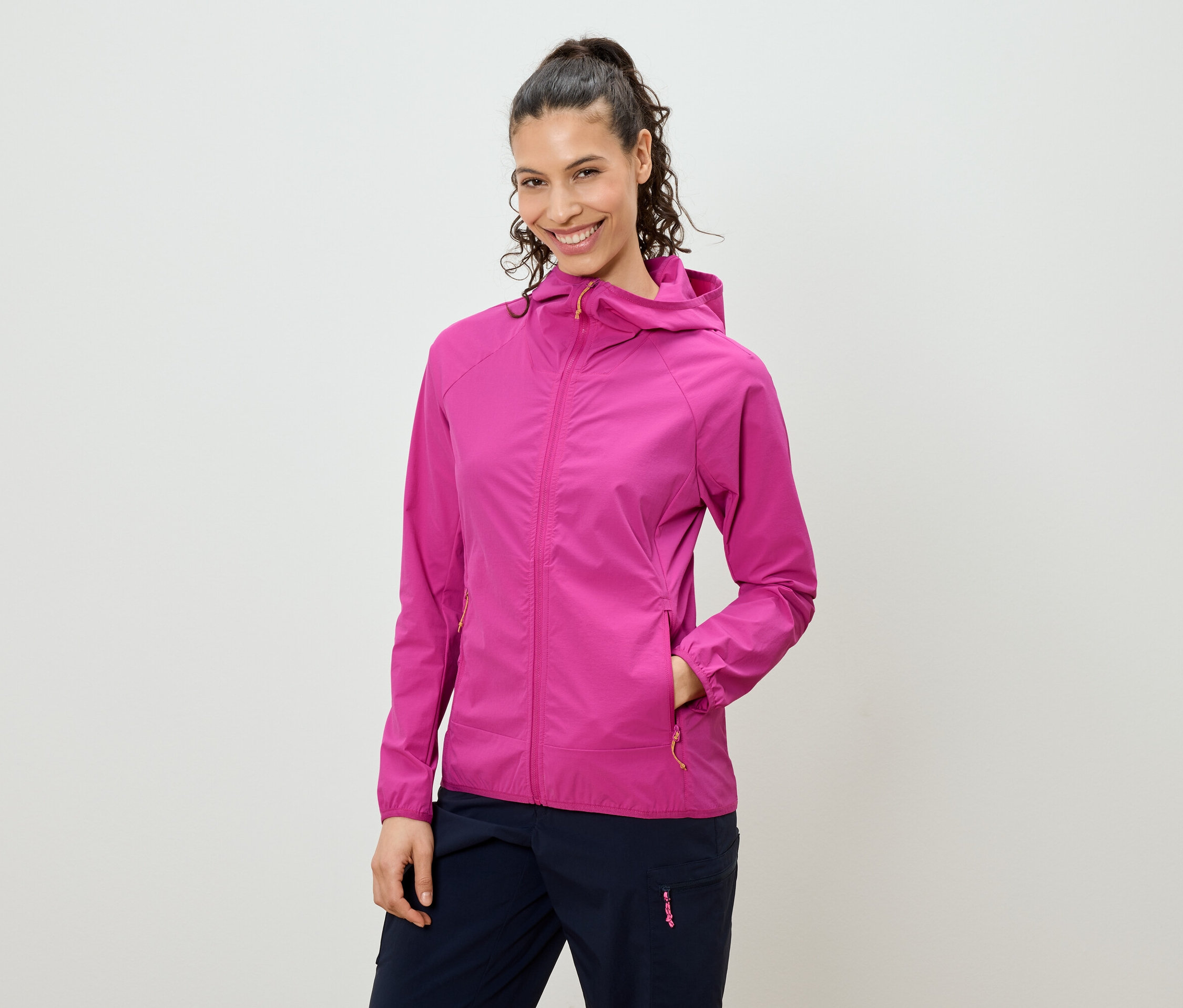 Mosolygó, copfba kötött hajú nő rózsaszín kapucnis kabátot és sötétkék nadrágot visel.