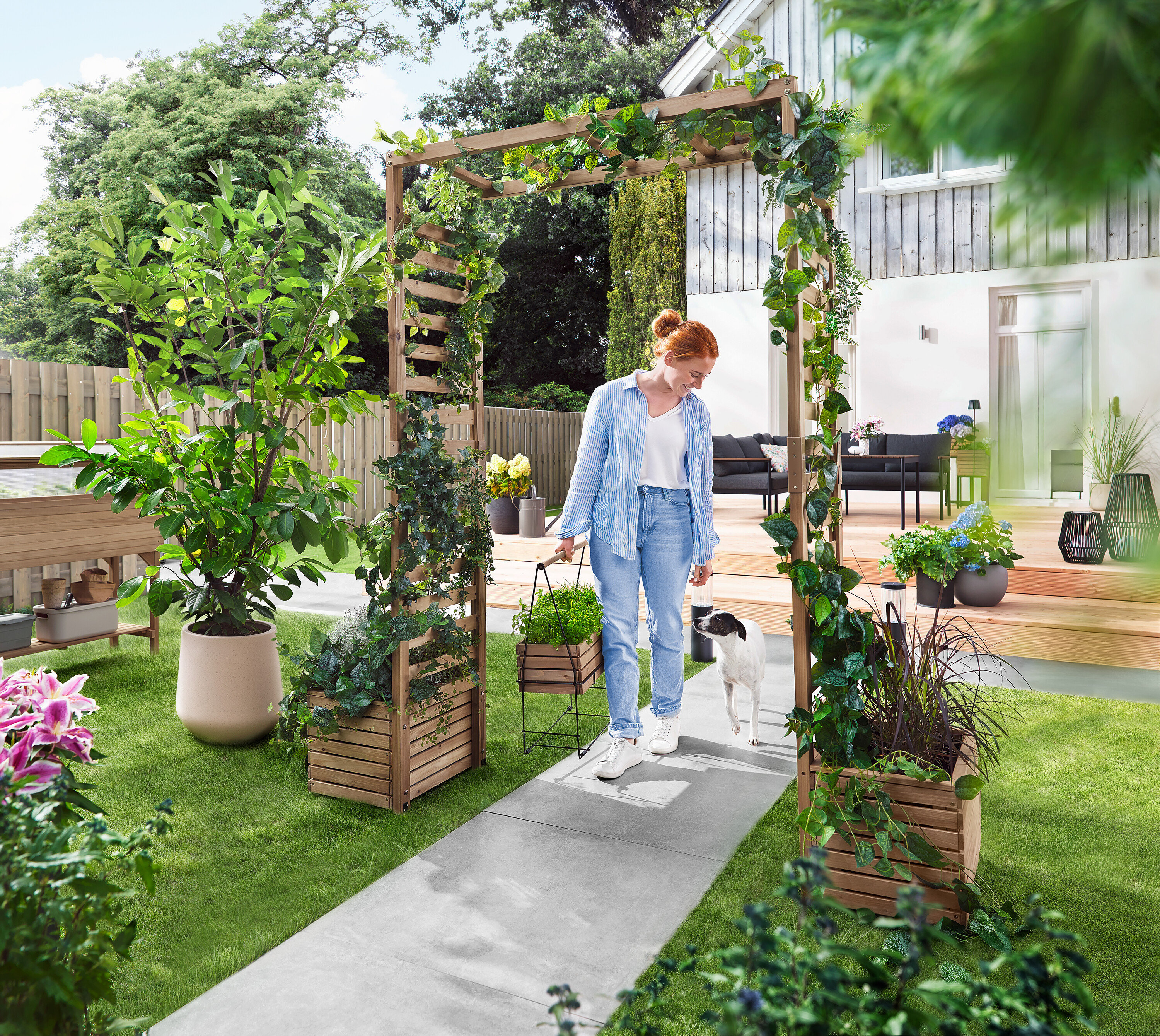 Egy nő egy kutyával sétál át egy növényekkel benőtt kerti boltíven. Egy virágládát visz.