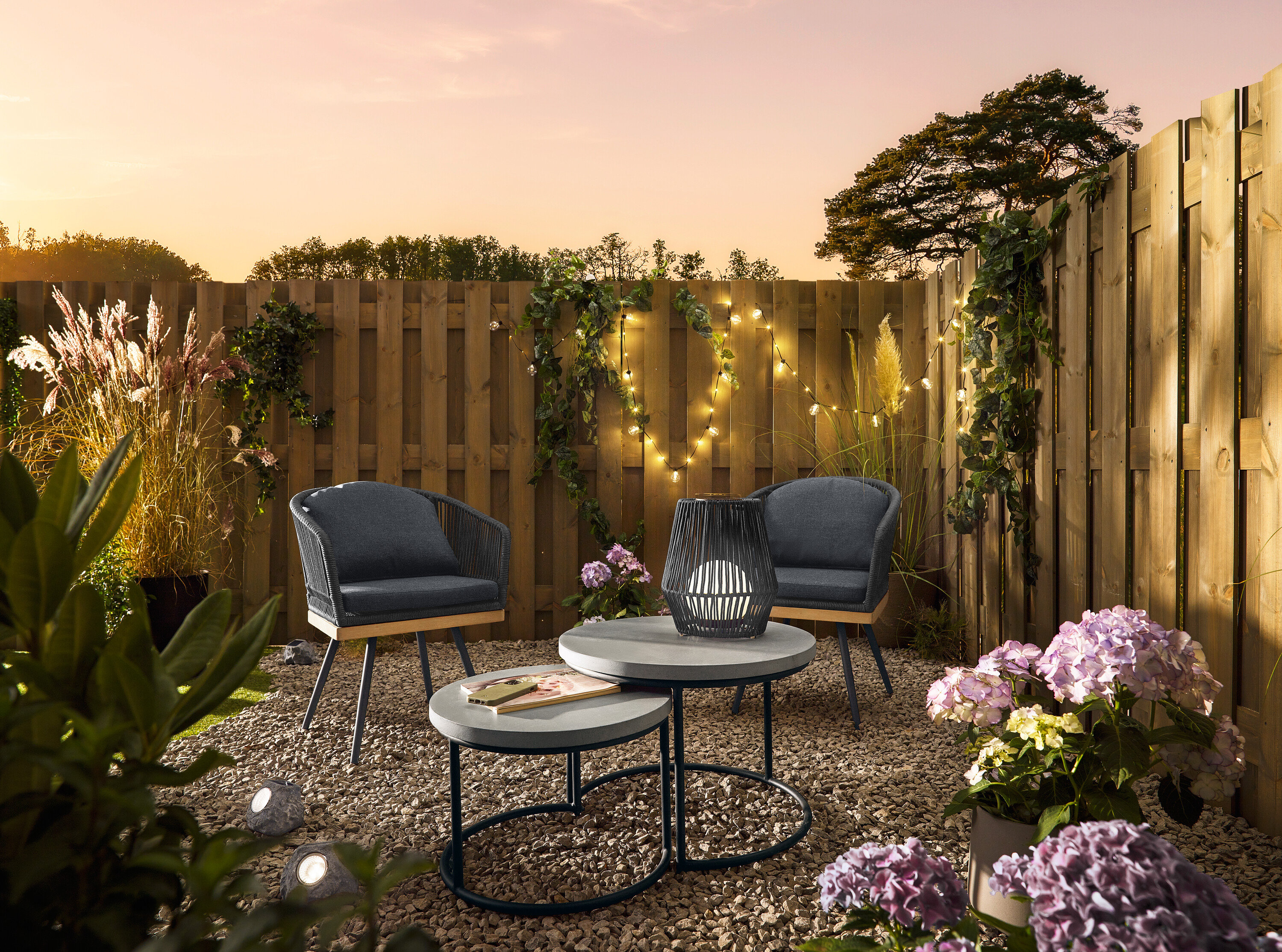 Két, textil keresztszőttel ellátott forgó kerti szék áll 2 db policement asztallapos kerti asztal mellett. Az egyiken egy kis, textilszalagos napelemes viharlámpa áll. Prémium napelemes LED-es kövek találhatók a kavicsban.