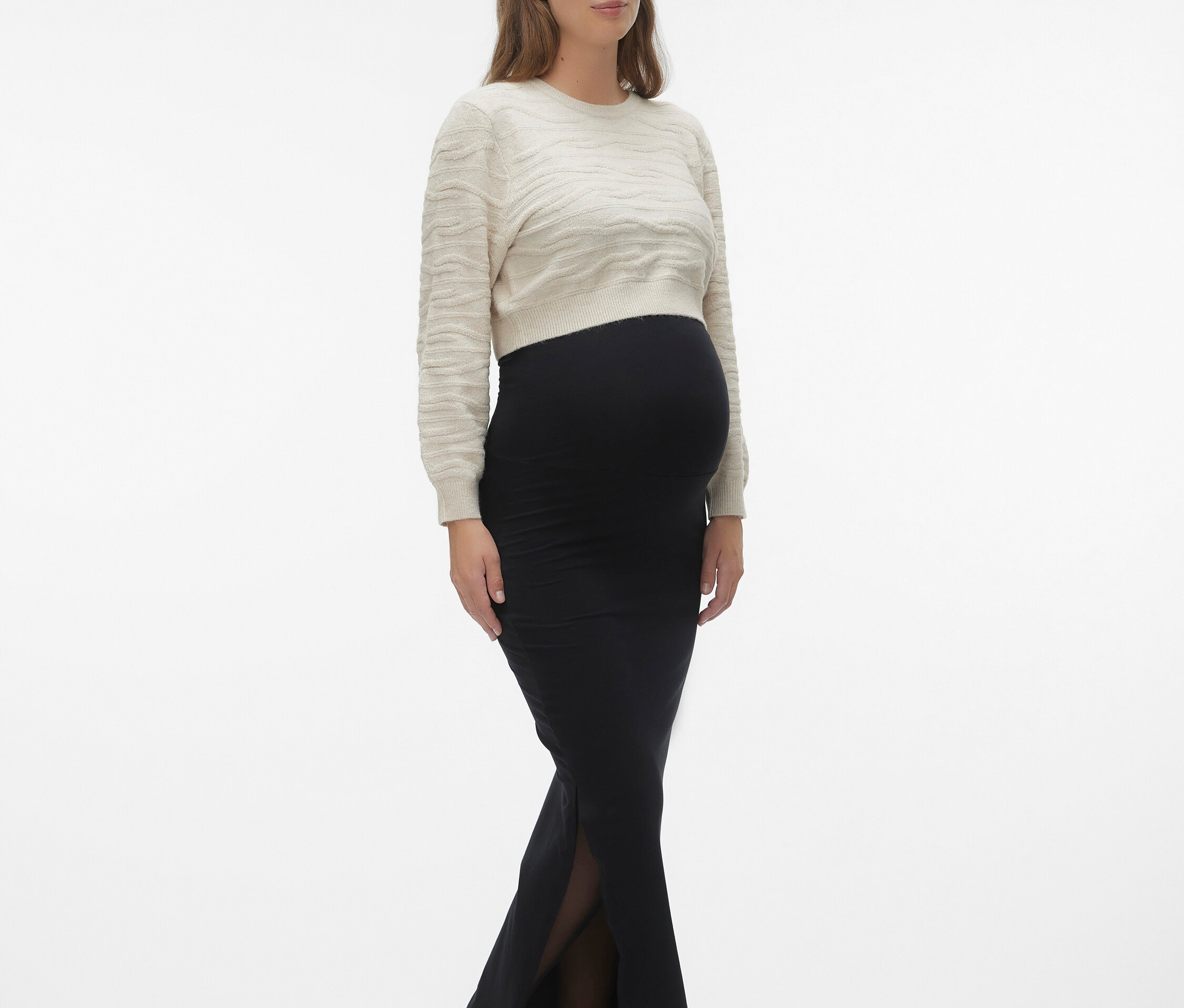 Terhes nő bézs színű pulóverben és hasítékkal ellátott fekete szoknyában, hozzá fekete cipő.