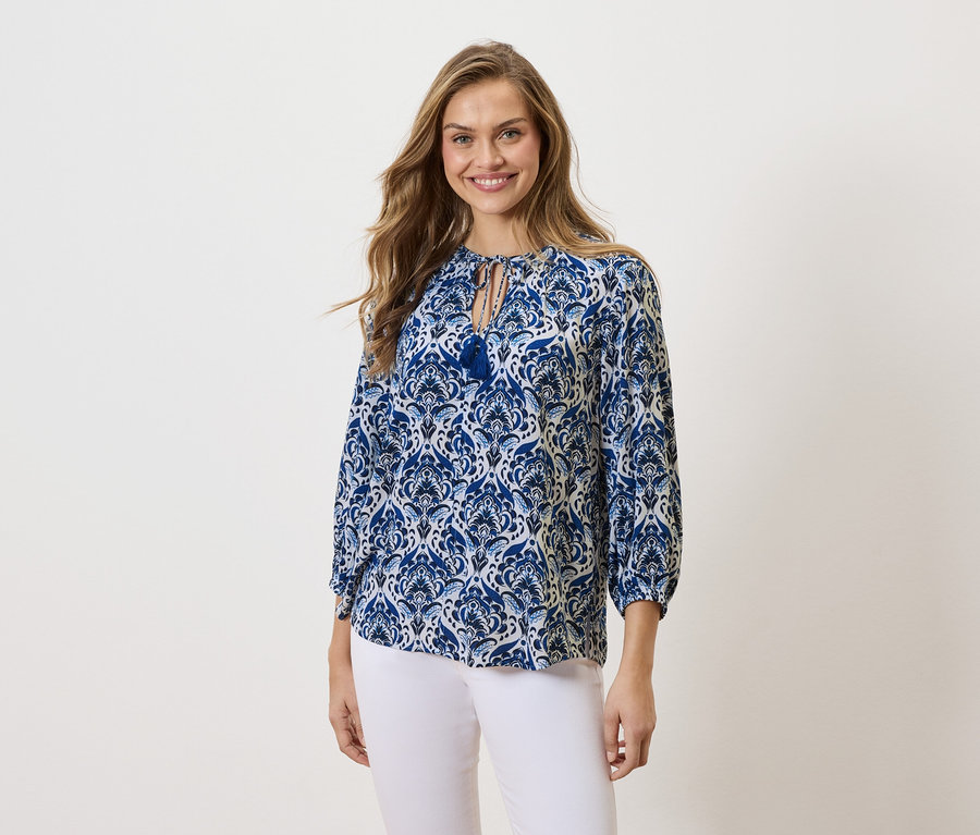 Hosszú szőke hajú nő mosolyog, kék-fehér mintás blúzt és fehér nadrágot visel.