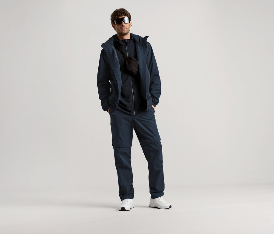 Férfi áll napszemüvegben, sötétkék kabátban, nadrágban és övtáskával világos háttér előtt.