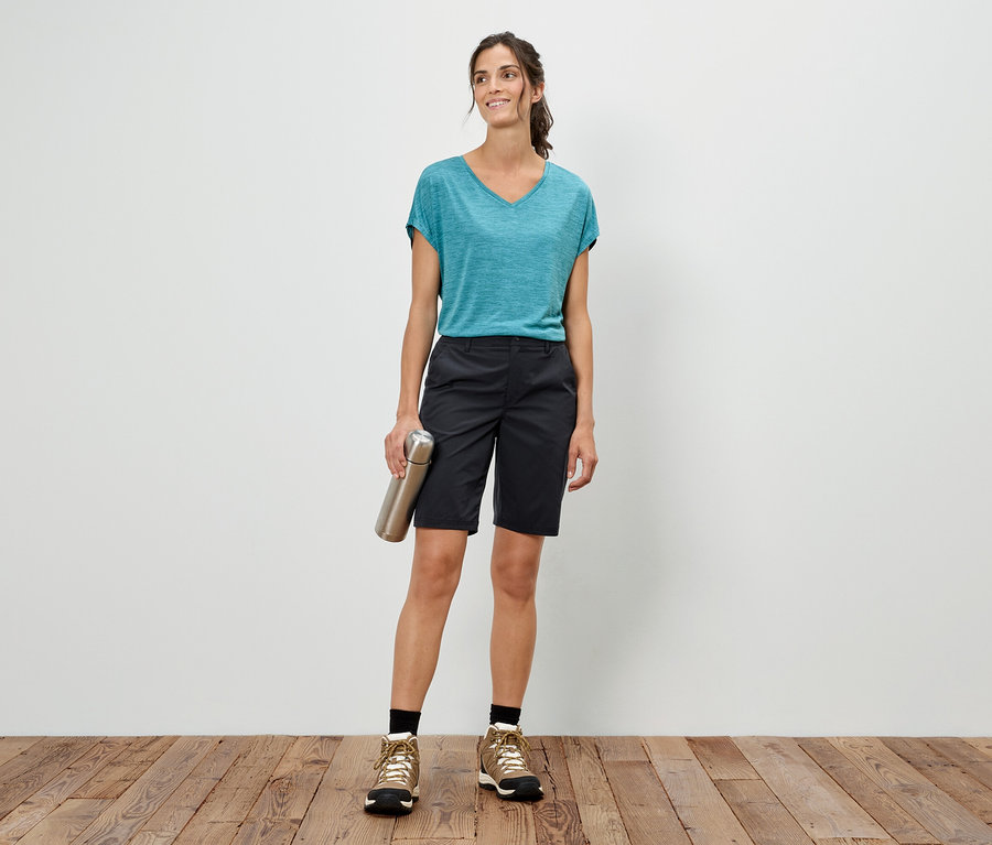 Nő áll egy fehér fal előtt, ezüst színű kulacsot tart, kék pólót, rövidnadrágot és túrabakancsot visel.