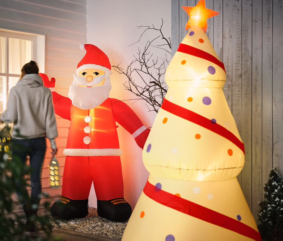 Egy nő sétál el egy XXL LED-es felfújható télapó és egy XXL LED-es felfújható karácsonyfa mellett.