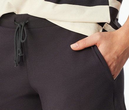 Egy személy közelről, sötétszürke SCHIESSER rövidnadrágos pizsamában, megkötött húzózsinórral és a jobb zsebében lévő kézzel. A rövidnadrág felett fekete-ekrü csíkos felső látható.