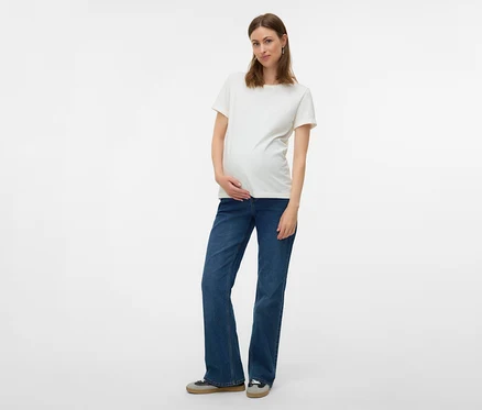 Terhes nő fehér pólóban és MAMALICIOUS kismama farmernadrágban pózol fehér háttér előtt.